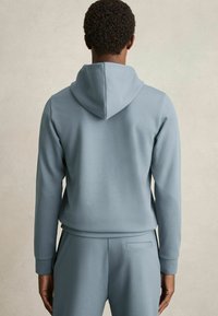 Felpa con cappuccio di colore azzurro chiaro, caratterizzata da una texture liscia, maniche lunghe e una vestibilità ampia, abbinata a pantaloni coordinati.
