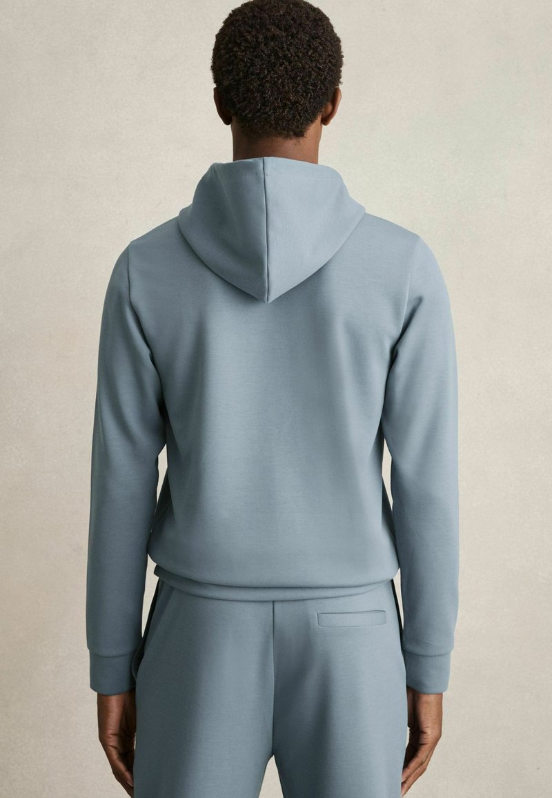 Felpa con cappuccio di colore azzurro chiaro, caratterizzata da una texture liscia, maniche lunghe e una vestibilità ampia, abbinata a pantaloni coordinati.