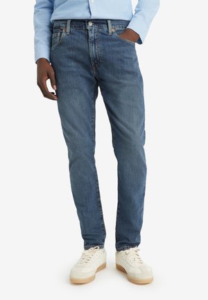 Homme portant un jean bleu, une chemise bleu clair et des baskets blanches, debout avec une main dans la poche sur un fond uni.