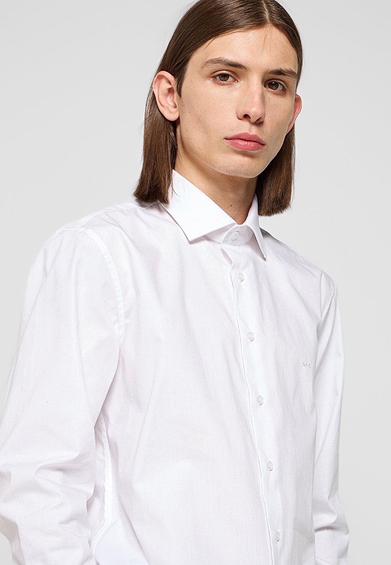 Junge Person mit schulterlangem braunem Haar, die ein weißes, knopfgeschlossenes Hemd trägt, vor einem einfarbigen Hintergrund.