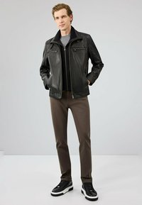 Zwarte leren jack met ritszakken, gestructureerde afwerking, gedragen over een grijze shirt en bruine broek, gecombineerd met zwarte sneakers.