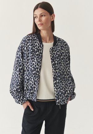 Woman wearing a navy and white spotted bomber jacket over a cream sweater and black pants, standing with hands in pockets, looking left.