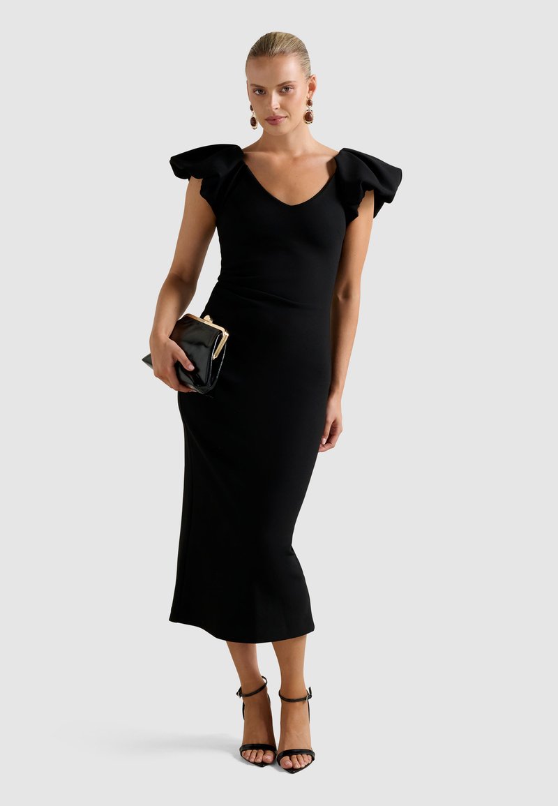 Vrouw in een zwart midi-jurk met gerimpelde mouwen, houdt een zwarte clutch vast, draagt zwarte enkelsandalen met bandjes en opvallende oorbellen.