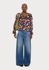 Blusa trasparente nera a fantasia floreale, vestibilità ampia, con maniche a sbuffo. Abbinata a jeans blu a gamba larga e a una piccola borsa verde oliva.