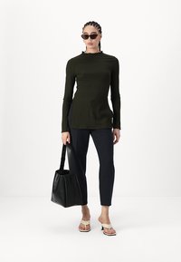 Olivegrünes Langarmshirt aus strukturiertem Stoff, kombiniert mit dunklen Hosen, beigen Sandalen und einer schwarzen Tragetasche. Stylisch ergänzt mit Sonnenbrillen.