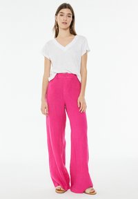 Woman wearing a white V-neck T-shirt tucked into bright pink high-waisted wide-leg pants with sandals, standing against a white background.
