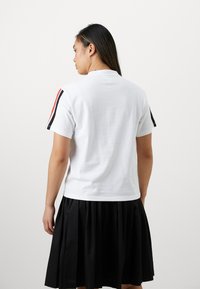 Λευκό βαμβακερό T-shirt με μαύρες και κόκκινες ρίγες στις μανσέτες, σε συνδυασμό με μια μαύρη πλάγια φούστα. Πίσω όψη, μινιμαλιστικός σχεδιασμός.