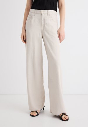 Nő, aki krémszínű, magas derekú, bőszárú nadrágot visel fekete ujjatlan felsővel és fekete, nyitott orrú, magassarkú szandállal.