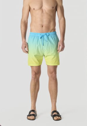 Hombre sin camiseta que lleva pantalones cortos de baño con un degradado de azul a amarillo y sandalias negras, de pie frente a un fondo liso.