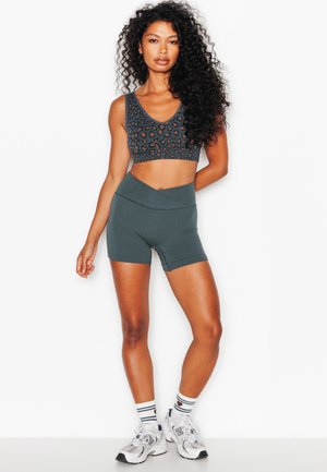 Vrouw met lang krullend haar die een sportbeha met luipaardprint, donkergroene biker shorts met hoge taille, witte sokken en witte sneakers draagt, staand.