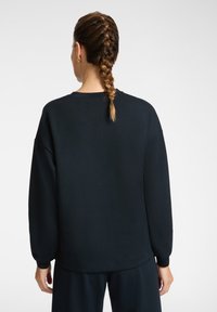 Donna con capelli intrecciati che indossa una felpa nera a maniche lunghe e pantaloni abbinati, vista di spalle su uno sfondo chiaro e semplice.