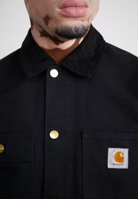 Giacca nera di cotone con colletto in corduroy, bottoni dorati e due tasche frontali. Presenta una piccola etichetta con il logo Carhartt arancione.