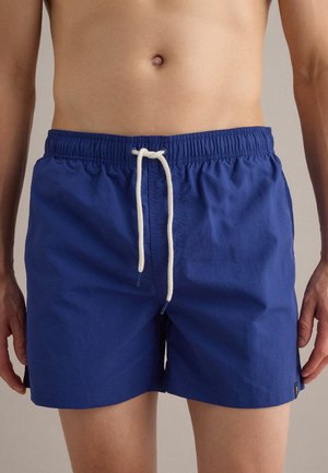 Torace maschile con pantaloncini da bagno blu navy a metà coscia con vita elastica e coulisse bianca, in piedi davanti a uno sfondo semplice.