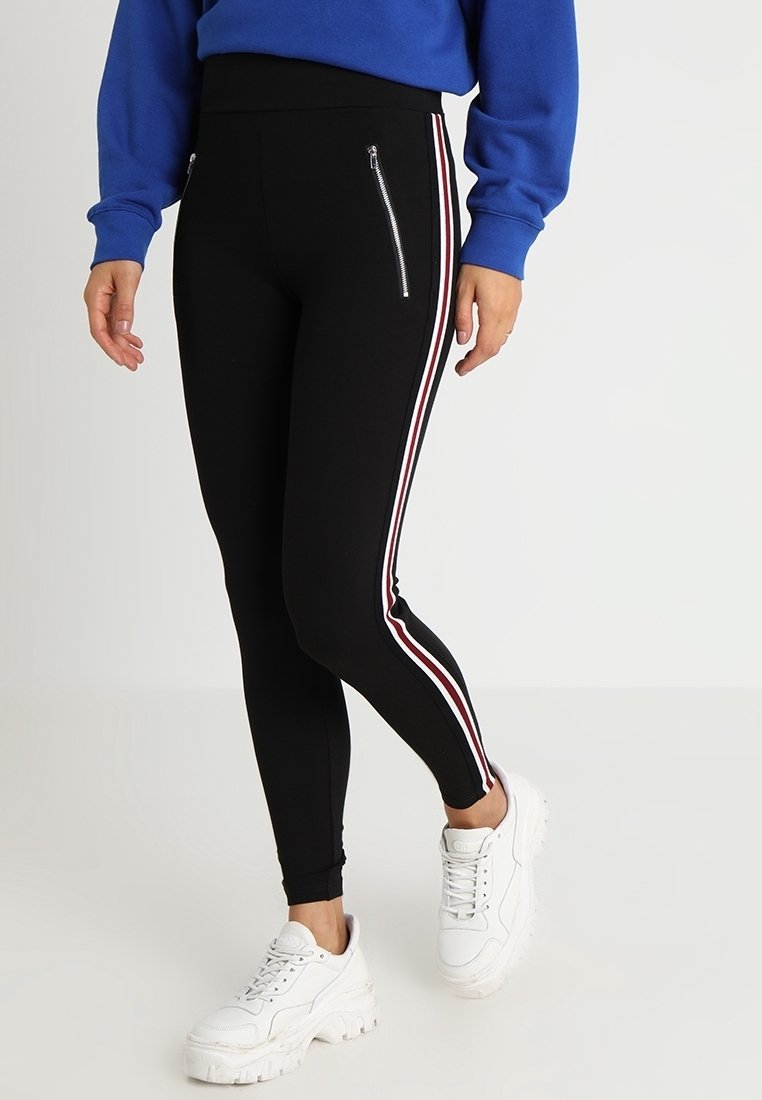 Leggings noirs avec des bandes latérales blanches et rouges, dotés de poches zippées et d'une taille haute, assortis à des baskets blanches.