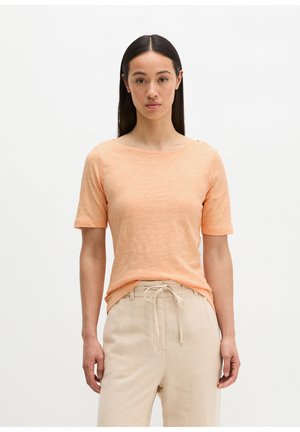 Frau mit langen dunklen Haaren, die ein pfirsichfarbenes kurzärmeliges Shirt und beigefarbene Hosen mit Kordelzug trägt, steht vor einem schlichten weißen Hintergrund.