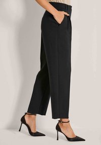 Vrouw in zwarte hoge, taps toelopende broek met handen in de zakken en zwarte spitse stilettohakken met enkelbandjes.