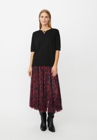 Kvinde iført en sort kortærmet sweater, rød og sort mønstret plisseret midi-nederdel, sorte støvler til midt på læggen og guld halskæde, stående.