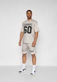 Light beige sports jersey and shorts set with black "CHIEFS" text, vertical stripes, and logo accents. Includes white trim and athletic shoes.
