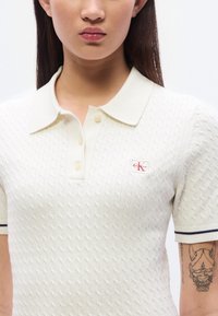 Κρεμ πόλο μπλουζάκι με πλέξη καλώδιο, με γιακά, που διαθέτει μικρό κόκκινο και λευκό λογότ υπο Calvin Klein και ριγωτές λεπτομέρειες στα μανίκια.