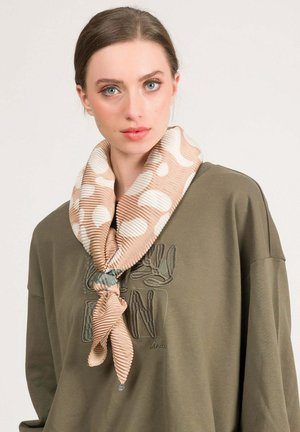 Giovane donna con occhi azzurri che indossa una sciarpa plissettata beige con motivi e una felpa verde oliva con testo ricamato.