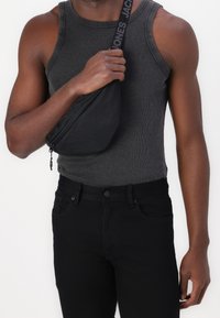 Black sling bag with zipper, worn across the shoulder, paired with a gray ribbed tank top and black jeans. Minimalistic design and texture.