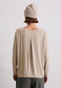 Beige langærmet top med rund halsudskæring og en plissédetalje på ryggen. Stoffet er glat, med en let figursyet pasform.