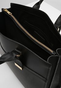 Svart läderhandväska med guldzipper och spänne, öppen för att visa inre fack och platta handtag på en vit yta.