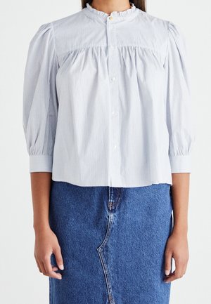 Kvinde iført en lyseblå skjorte med striber og puffede tre-kvart ærmer samt en blå denim nederdel med asymmetrisk søm.