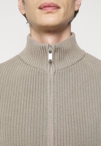 Texturerad beige stickad jacka med hög krage och full dragkedja, med ett fint ribbat mönster och släta dragkedjespännen.