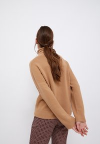 Pull col roulé kaki à coupe décontractée, avec poignets et ourlet côtelés, assorti à un pantalon marron à motifs petits dessins géométriques.