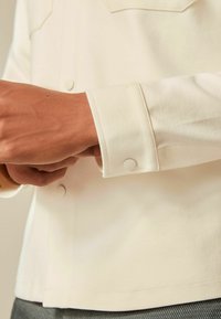 Cremefarvet skjorte med tekstureret stof, der har en lukket ærme og rund kant. Ærmet har en subtil knaplukningsdetalje.
