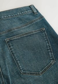 Gros plan sur la zone de la poche arrière d'un jean en denim bleu avec des coutures visibles et une passants de ceinture sur un fond neutre.