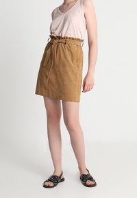 Brun mockaskjol med bälte i midjan och två framfickor, parat med en ljus tanktopp och prickiga svarta sandaler.