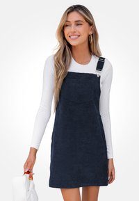 Vestido peto de pana en color navy con tirantes ajustables, bolsillo frontal y un corte relajado, combinado con una blusa blanca de manga larga ajustada.
