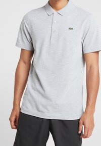 Grå pikétröja i bomullsblandning. Har en klassisk krage, treknäppning och en liten grön broderad logotyp på bröstet.