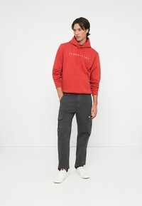 Červená mikina s predným vreckom a bielym textom, v kombinácii s tmavými cargo nohavicami s bočnými vreckami a bielymi teniskami. Jednoduchý, neformálny outfit.