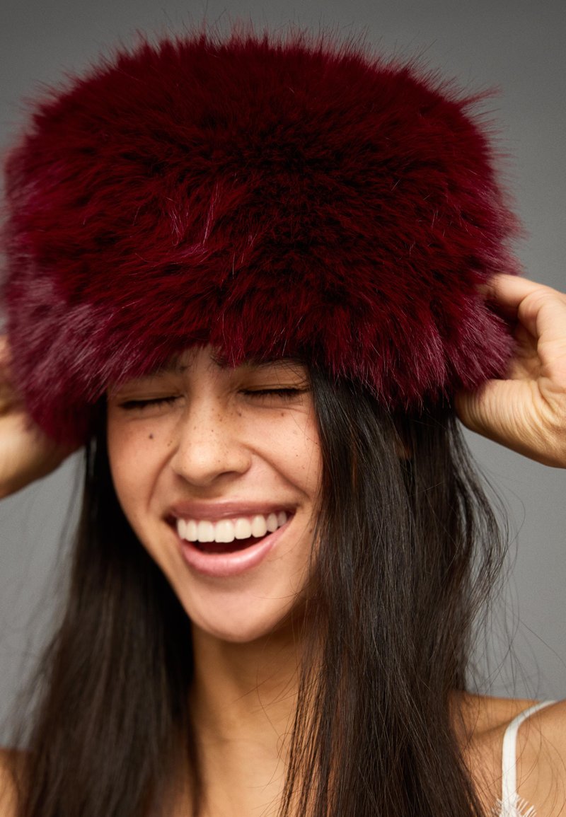 Șapcă din blană falsă de culoare burgundă, cu un design voluminos și pufos. Textura este moale și groasă, oferind căldură și acoperire.