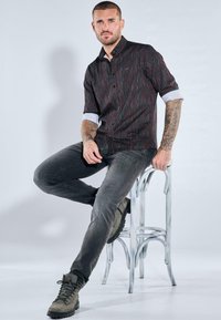 Schwarz-rot gestreiftes Hemd mit Knöpfen und hochgekrempelten Ärmeln, kombiniert mit dunkelgrauen Jeans und olivgrünen Schnürstiefeln, auf einem Metallhocker sitzend.