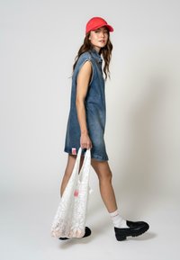Dongerikjole uten ermer, lyseblå, med logo-patch. Svarte klumpete sko og en hvit lace tote bag. Rød caps og hvite ankelstrømper.
