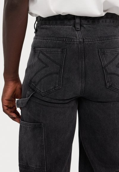 Melnas denim cargo bikses ar brīvu piegriezumu, kurām ir lielas aizmugurējās kabatas, izšūtas akcenti un izturīga tekstūra.
