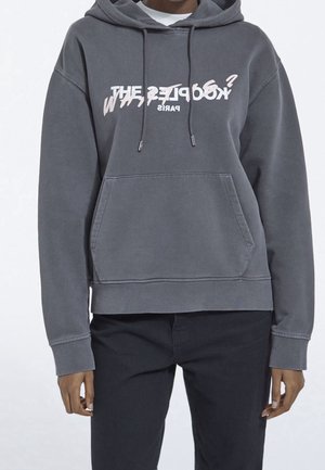 Persoon draagt een vervaagde grijze capuchontrui met voorzak en een gelaagd wit en vervormd tekstontwerp op de borst, gecombineerd met een zwarte broek.