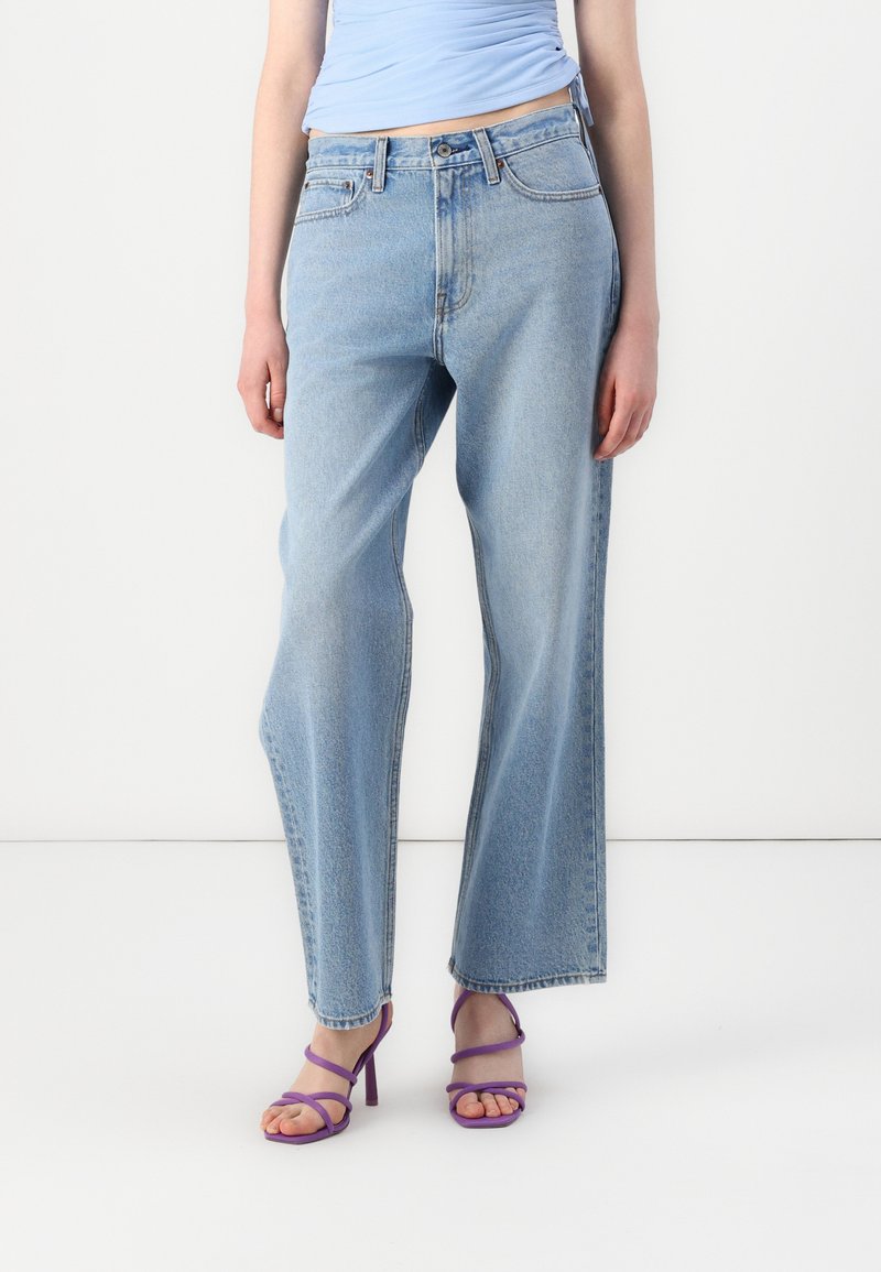 Jeans de pierna ancha de color azul claro, hechos de denim, con una cintura alta y una textura ligeramente desvanecida. Combinados con tacones morados con tiras.