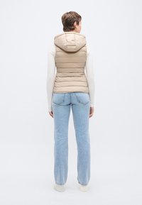 Μπεζ μπουφάν με επένδυση και κουκούλα, με κάθετες ραφές και εφαρμοστή γραμμή, συνδυασμένο με ανοιχτόχρωμο τζιν και λευκά αθλητικά παπούτσια.