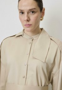Blusa beige de botones con cuello estructurado y una solapa en el hombro adjunta. Textura suave con botones a juego.