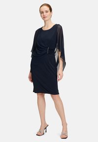 Granatowa sukienka z lekko opadającymi, przejrzystymi rękawami w kształcie peleryny, zebranym detalem w talii z metalicznym akcentem, do kolan, wykonana z gładkiego materiału. Czarne sandały na obcasie.