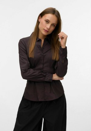 Femme aux longs cheveux bruns portant une chemise foncée ajustée boutonnée et un pantalon noir, posant avec une main près de son visage sur un fond blanc.