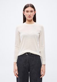 Pulover bej deschis cu mâneci lungi, textură moale, cu guler rotund, combinat cu pantaloni înalți, de culoare gri închis, cu o curea neagră și detalii subtile.