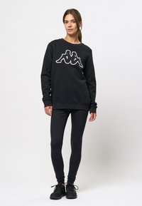 Zwarte sweatshirt met witte Kappa-logo, geribbelde ronde hals, en strakke zwarte legging. Model draagt zwarte sportschoenen.