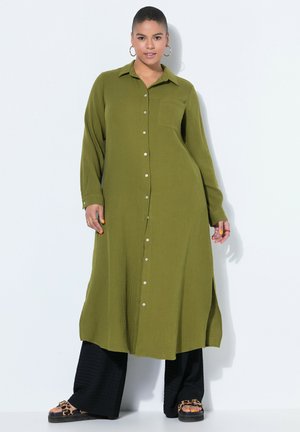 Donna che indossa un lungo vestito oliva con bottoni sopra pantaloni neri a gamba larga e sandali piattaforma con stampa leopardata, in piedi su sfondo bianco.