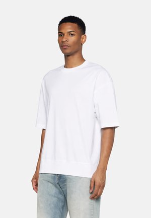 Joven con una camiseta blanca lisa de manga corta y oversize y vaqueros azul claro, de pie contra un fondo liso.